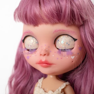 Pode incluir: Um close-up de uma boneca com cabelo rosa e tez clara. A boneca tem olhos grandes com glitter e delineador preto. As bochechas da boneca têm detalhes roxos e brancos. A boneca está vestindo um vestido creme.