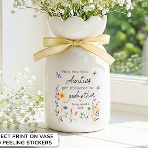 May include: White ceramic vase with a scalloped top, tied with a gold ribbon, filled with white flowers. The vase has text that reads "Only the best Aunties get promoted to godmother. Love, Annie 2026" with floral and butterfly accents.