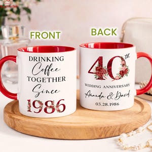 May include: Two white ceramic mugs with red interiors and handles. The left mug features the text "DRINKING Coffee TOGETHER Since 1986" with floral accents. The right mug displays "40th WEDDING ANNIVERSARY Amanda & David 03.28.1986" with floral details.
