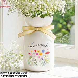 May include: A white ceramic vase with a scalloped top and a gold ribbon tied around the neck. The vase is decorated with a colourful floral design and the text "Add your message here". The vase is filled with white flowers.
