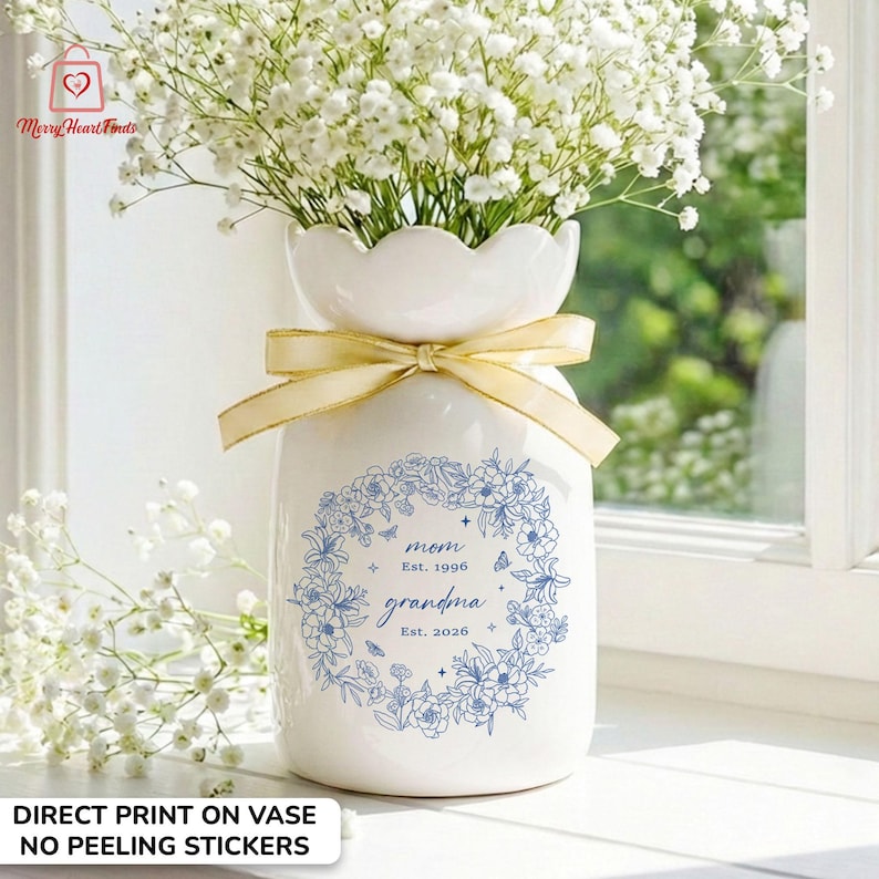 Puede incluir: Un jarr&oacute;n de cer&aacute;mica blanco con una parte superior festoneada, lleno de flores blancas y atado con una cinta dorada. El jarr&oacute;n tiene un dise&ntilde;o de corona floral azul con las palabras "mom Est. 1996 grandma Est. 2026". El texto "DIRECT PRINT ON VASE NO PEELING STICKERS" tambi&eacute;n es visible.