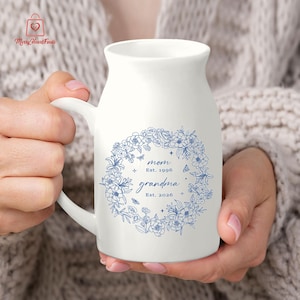 Puede incluir: Taza de cer&aacute;mica blanca con asa, adornada con una corona floral azul. La taza tiene la inscripci&oacute;n "mom Est. 1996 grandma Est. 2026". El logotipo de Merry Heart Finds est&aacute; en la esquina superior izquierda. La taza es sostenida.