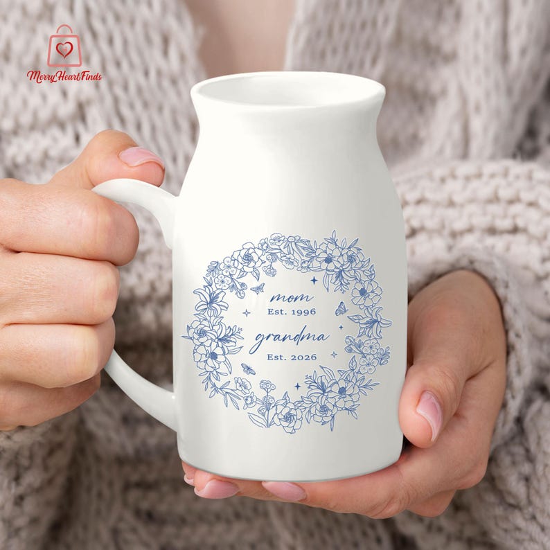 Puede incluir: Taza de cer&aacute;mica blanca con asa, adornada con una corona floral azul. La taza tiene la inscripci&oacute;n "mom Est. 1996 grandma Est. 2026". El logotipo de Merry Heart Finds est&aacute; en la esquina superior izquierda. La taza es sostenida.