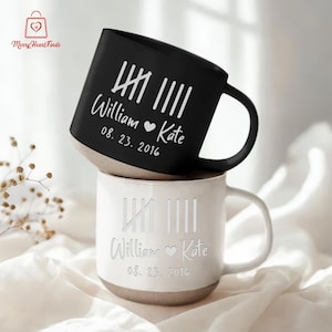 May include: Two ceramic mugs, one black and one white, stacked on top of each other. The black mug has white text that reads "William ♥ Kate" and "08. 23. 2016". The white mug has the same text. Both mugs have a light brown base.