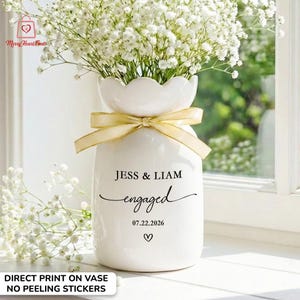 May include: A white ceramic vase with a scalloped rim, tied with a gold ribbon, and filled with white flowers. The vase has the words "JESS & LIAM engaged 07.22.2026" printed on it, with a heart symbol below. The text "DIRECT PRINT ON VASE NO PEELING STICKERS" is also visible.