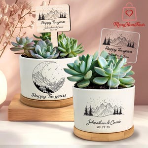 May include: White ceramic succulent pots with a mountain and moon design, and wooden signs that read "Happy Tin years" with a date and names. The pots sit on bamboo saucers, and contain green succulents.