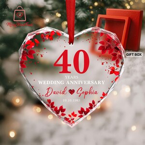 May include: A heart-shaped glass ornament with red detailing and a red ribbon. The ornament celebrates a 40th wedding anniversary, with the names "David & Sophia" and the date "10.10.1985" displayed. A red gift box is visible in the background.