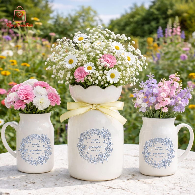Puede incluir: Tres jarrones de cer&aacute;mica blanca con arreglos florales. Los jarrones est&aacute;n decorados con una corona floral azul y el texto "mom est. 1998 grandma est. 2016". Las flores incluyen margaritas, claveles y otras flores coloridas.