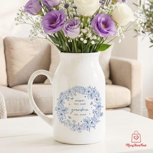 Puede incluir: Una jarra de cer&aacute;mica blanca con asa, adornada con un dise&ntilde;o de corona floral azul y las palabras "mom Est. 1996 grandma Est. 2026". La jarra contiene un ramo de flores moradas y blancas. Un regalo perfecto.