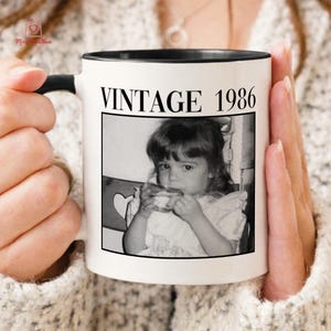 May include: White ceramic mug with a black interior and handle. The mug features the text "VINTAGE 1986" above a black and white photo of a young child. The mug is held by a person wearing a cream-colored sweater.
