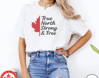 May include: A white short-sleeved t-shirt featuring a red maple leaf graphic next to the black text True North Strong & Free. The shirt is paired with blue denim jeans that have distressed detailing on the front of the legs.