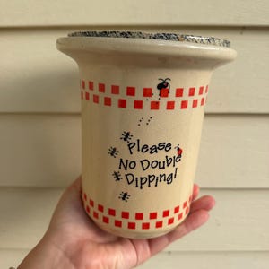 May include: A cream-colored ceramic crock featuring a red checkered border at the top and bottom. The center displays the black text "Please No Double Dipping!" surrounded by small illustrations of ants and a ladybug on a speckled gray lid.