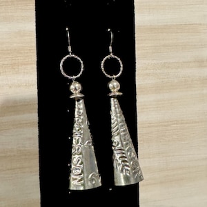 May include: A pair of silver-toned dangle earrings. Each earring features a cone-shaped pendant with embossed floral and text details, suspended from a small ring and bead. The text on the earrings reads "Jefferson".