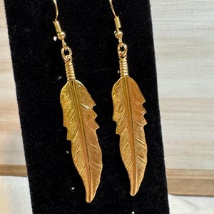 May include: A pair of gold-coloured feather earrings. Each earring features a detailed feather design with a textured surface and a decorative hook. The earrings are displayed against a black background.