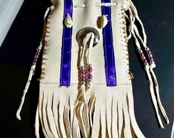 May include: Cream-colored leather pouch with purple ribbon accents and fringe. The pouch features a silver-colored button closure and is adorned with small beads and shell-like embellishments. The design is reminiscent of Native American crafts.