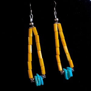 May include: A pair of handcrafted earrings featuring a triangular design. The earrings are composed of yellow cylindrical beads, silver accents, and turquoise stone accents. The earrings hang from silver hooks against a black background.