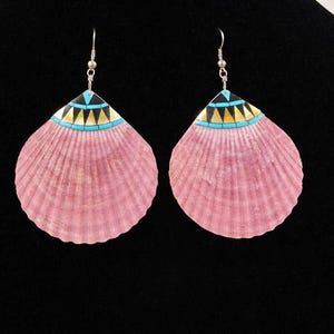 May include: A pair of pink shell earrings with a decorative top featuring black, yellow, and turquoise geometric patterns. The earrings hang from silver-colored hooks, showcasing a unique, handcrafted design.