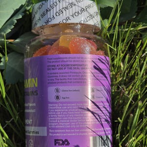 Puede incluir: Una botella de pl&aacute;stico transparente llena de vitaminas gomosas de colores, colocada en un lecho de hierba verde. La botella tiene una tapa blanca y una etiqueta morada con texto. La etiqueta indica que el producto no contiene gluten ni huevo.