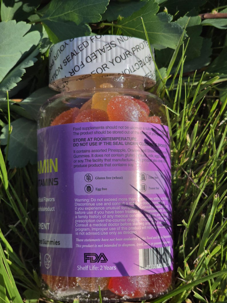 Puede incluir: Una botella de pl&aacute;stico transparente llena de vitaminas gomosas de colores, colocada en un lecho de hierba verde. La botella tiene una tapa blanca y una etiqueta morada con texto. La etiqueta indica que el producto no contiene gluten ni huevo.