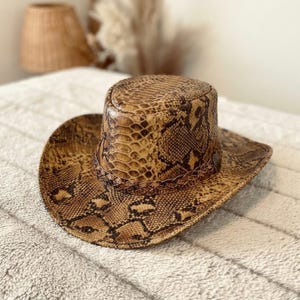 May include: A cowboy hat featuring a brown and tan snakeskin pattern throughout the crown and brim. The hat includes a braided band encircling the base of the crown, which is finished with a small, metallic heart-shaped accent on the side.