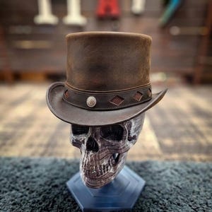 May include: A brown leather top hat featuring a structured crown and a curved brim. The hat band is adorned with a series of diamond-shaped cutouts and a central circular silver-toned metal emblem, resting upon a gray and white skull display stand.