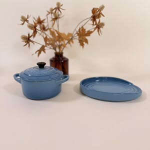 Puede incluir: Una olla holandesa Le Creuset azul claro con un pomo negro y una fuente para servir a juego. La olla holandesa tiene dos asas. Un jarrón de flores secas está en el fondo.