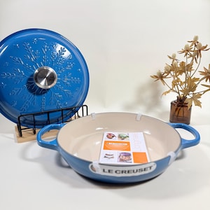 May include: A blue Le Creuset Dutch oven with a matching lid. The lid features a snowflake design and a silver handle. The oven has a cream-colored interior and blue handles. A small brochure is inside the oven.