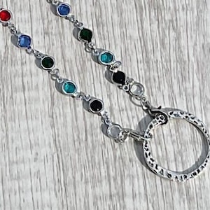 May include: A silver-toned keychain with two strands of colorful, round gemstones. The gemstones are red, blue, green, teal, black, and clear. The keychain has a large, patterned ring at the end.