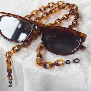 May include: Sunglasses with a tortoiseshell pattern frame and dark lenses, paired with a matching chain. The chain is made of interlocking links and has rubber ends for attaching to the glasses. The sunglasses rest on a white surface.