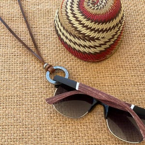May include: Sunglasses with brown patterned arms and dark lenses, paired with a leather eyeglass holder. A woven basket with red, brown, and tan patterns sits nearby. The items are arranged on a textured, light brown surface.