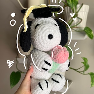 May include: A handmade white and black Snoopy plushie wearing a graduation cap and holding a pink and green flower. The plushie has black eyes and black accents on its paws. The graduation cap is black with a yellow tassel.