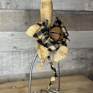 May include: A rustic potato masher with a wooden handle and a metal mashing head. It is adorned with a tan and black plaid fabric bow and a brown bell. The masher is set against a wooden background.