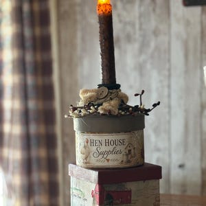May include: A decorative candle with an orange flame sits atop two stacked boxes. The top box reads "Hen House Supplies" and features a chicken illustration. The bottom box has "Farm Fresh" and "Eggs" printed on it.