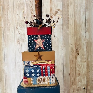 May include: A decorative patriotic candle holder with a faux candle and stacked boxes. The boxes are red, white, and blue, decorated with stars, stripes, and the American flag. The candle has a flickering orange light.