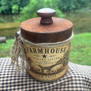 May include: A cylindrical container with a rusted metal lid and a dark wooden knob. The label features the text FARMHOUSE COUNTRY LIVING Simple Blessings FARM FRESH, with illustrations of pigs and a barn. Jute twine wraps around the top edge.