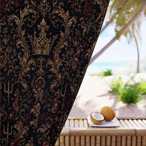 May include: A dark curtain featuring an intricate gold and red damask pattern with crown and trident motifs. The fabric hangs beside a bamboo surface holding a halved coconut on a white plate, with a blurred tropical beach view in the background.