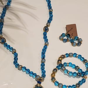 Pode incluir: Um colar de contas de vidro azul com um pingente envolto em ouro, um bracelete a condizer de contas de vidro azul e brincos. As contas têm um padrão de espiral azul claro e azul escuro.