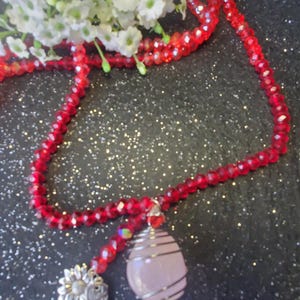 May include: A red crystal beaded necklace with a pink rose quartz pendant wrapped in silver wire. The pendant is accented with a silver charm featuring a flower design.