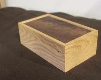 May include: A rectangular wooden box with a contrasting dark brown top and light brown sides. The box is made of wood and has a simple, clean design. The wood grain is visible on the sides.