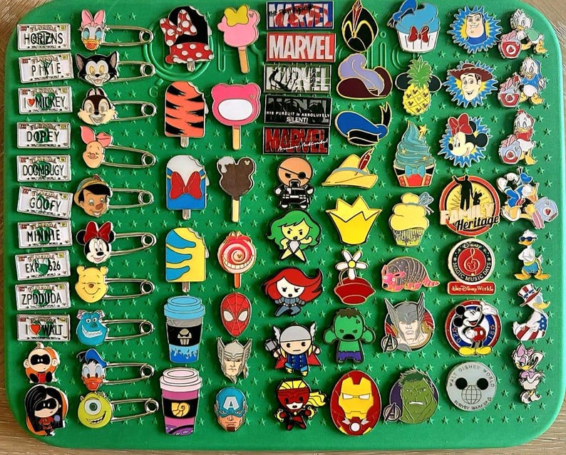 May include: A collection of colorful enamel pins featuring various Disney and Marvel characters, ice cream treats, and license plate designs. The pins are attached to a green pin board with safety pins.