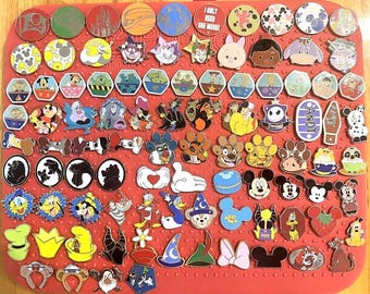 May include: A collection of Disney character pins, including Mickey Mouse, Donald Duck, and other popular characters. The pins are in various shapes and colors, arranged on a red surface. Some pins feature text like "I only need one more!"