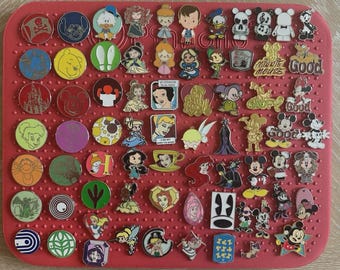 Könnte beinhalten: Eine Sammlung bunter Emaille-Anstecknadeln mit verschiedenen Disney-Charakteren und -Designs. Die Anstecknadeln sind auf einer roten, strukturierten Oberfläche angeordnet. Zu den Charakteren gehören Micky Maus, Winnie Puuh und Disney-Prinzessinnen.