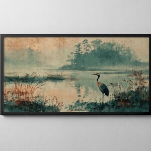Op de afbeelding: Een rechthoekige wandkunstprint in een zwarte lijst met een landschapsschilderij van een kraanvogel in een moerassig gebied. De scène bevat gedempte blauwgroene en perzikkleurige tinten, hoog gras en een verre bomenrij in het water.