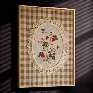 Op de afbeelding: Een ingelijst wandkunstwerk met een wit stoffen ovaal gecentreerd op een saliegroene en witte gingham-achtergrond. Het ovaal is omrand met cr&egrave;mekleurig kant en toont een geborduurde botanische illustratie van rode aardbeien en groene bladeren.