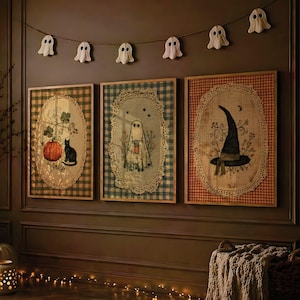 May include: Three framed rectangular wall art pieces featuring oval burlap centers with lace borders. The left print shows a pumpkin and black cat, the center a ghost, and the right a witch hat. A string of six small white ghost ornaments hangs above.