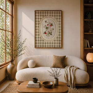 Op de afbeelding: Een ingelijst wandkunstwerk met een wit stoffen ovaal met geborduurde rode aardbeien en groene bladeren, omringd door een cr&egrave;mekleurige kanten rand, tegen een beige en wit ruitjesachtergrond in een dunne houten lijst.