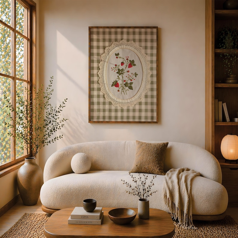 Op de afbeelding: Een ingelijst wandkunstwerk met een wit stoffen ovaal met geborduurde rode aardbeien en groene bladeren, omringd door een cr&egrave;mekleurige kanten rand, tegen een beige en wit ruitjesachtergrond in een dunne houten lijst.