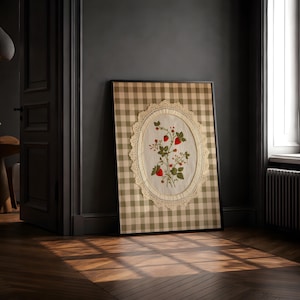 Op de afbeelding: Een ingelijst rechthoekig wandkunstwerk met een beige en witte gingham-achtergrond. In het midden bevindt zich een ovaal, cr&egrave;mekleurig stoffen paneel met een kanten rand, voorzien van een geborduurd botanisch ontwerp met rode aardbeien.