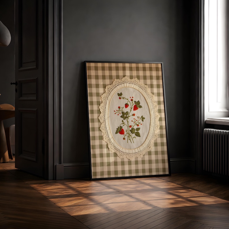 Op de afbeelding: Een ingelijst rechthoekig wandkunstwerk met een beige en witte gingham-achtergrond. In het midden bevindt zich een ovaal, cr&egrave;mekleurig stoffen paneel met een kanten rand, voorzien van een geborduurd botanisch ontwerp met rode aardbeien.