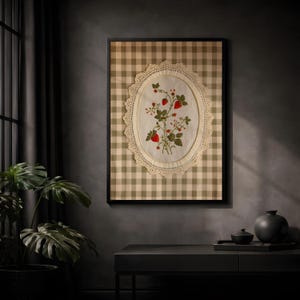 Op de afbeelding: Een ingelijst kunstwerk met een beige en witte gingham-achtergrond. In het midden bevindt zich een ovaal stoffen paneel met een delicate cr&egrave;mekleurige kanten rand, voorzien van een geborduurde botanische illustratie van rode aardbeien.